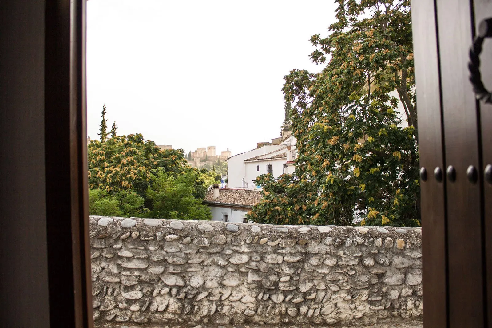 The house door open onto a stone wall, with neighbourhood houses and the Alhambra in the distance among the trees